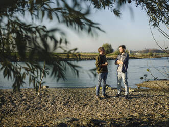 Son and father talking with each other at riverbank on sunny day - GUSF05191