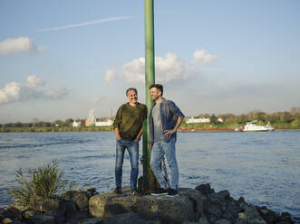 Smiling son and father leaning by pole while standing on rock against river - GUSF05193