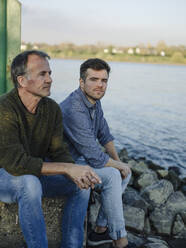 Father and son contemplating while sitting on rock against river - GUSF05194