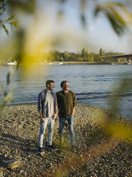 Father with arm around son against river on sunny day - GUSF05199