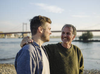 Happy father with arm around son at riverbank on sunny day - GUSF05200