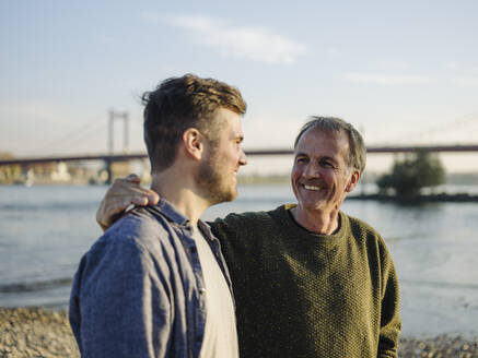 Glücklicher Vater mit Arm um Sohn am Flussufer an einem sonnigen Tag - GUSF05200