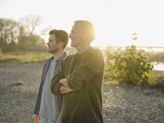 Father with arms crossed standing by son on riverbank at evening - GUSF05215