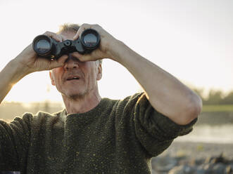 Senior man looking through binoculars against clear sky at riverbank - GUSF05224