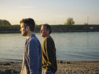 Father and son looking away while spending leisure time by river at evening - GUSF05228