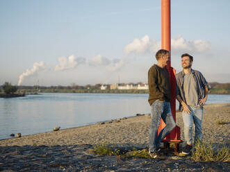 Smiling son and father leaning by pole while talking with each other at riverbank - GUSF05229