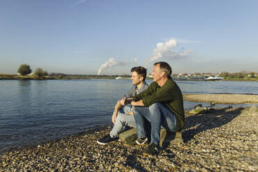 Father and son admiring river while sitting on rock during sunny day - GUSF05235