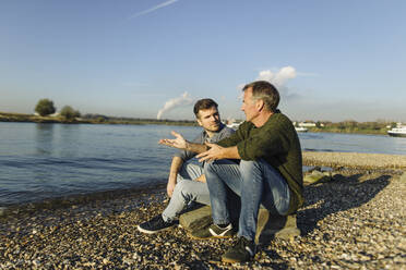 Father giving advice to son while sitting on rock at riverbank during sunny day - GUSF05236