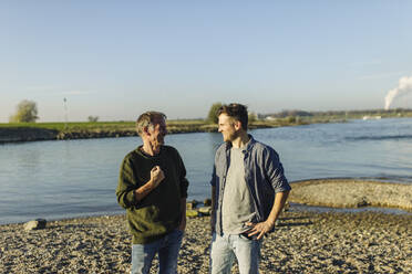 Cheerful father and son discussing while standing against river on sunny day - GUSF05237