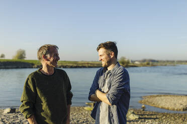 Glücklicher Sohn im Gespräch mit Vater am Fluss an einem sonnigen Tag - GUSF05238