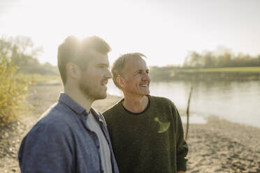 Sohn und Vater schauen an einem sonnigen Tag am Flussufer weg - GUSF05241