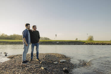 Son and father looking away while standing against clear sky at riverbank - GUSF05246