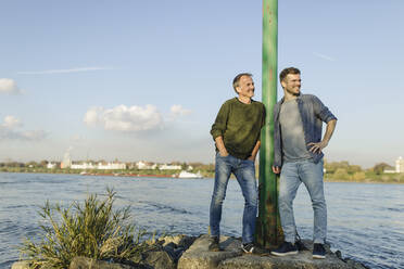 Smiling son and father leaning by pole while looking away at riverbank against sky - GUSF05250