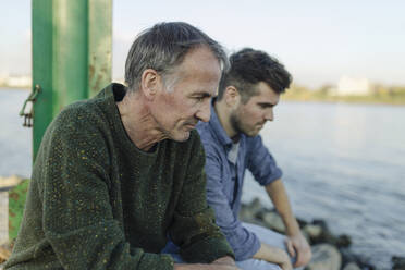 Son and father looking down while sitting by river - GUSF05261