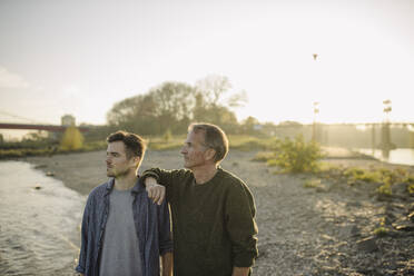 Father and son looking away while standing on riverbank at evening - GUSF05272