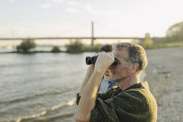 Vater schaut durch ein Fernglas und steht mit seinem Sohn am abendlichen Flussufer - GUSF05281