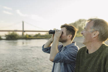 Son looking through binoculars while father laughing by on riverbank at evening - GUSF05284