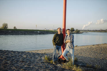 Thoughtful father and son leaning by pole against sky at riverbank - GUSF05286