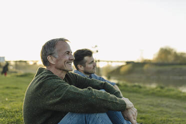 Smiling father spending leisure time with son on grass at riverbank - GUSF05287