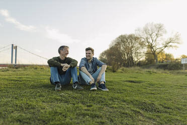 Glücklicher Vater und Sohn unterhalten sich im Gras sitzend gegen den Himmel - GUSF05288