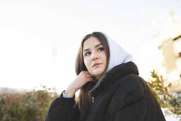 Young woman wearing hood clothing looking away while standing against sky - JCCMF01064