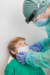 Side view of medic in protective uniform conducting PCR test on COVID virus while taking nasal swab from kid - ADSF20521