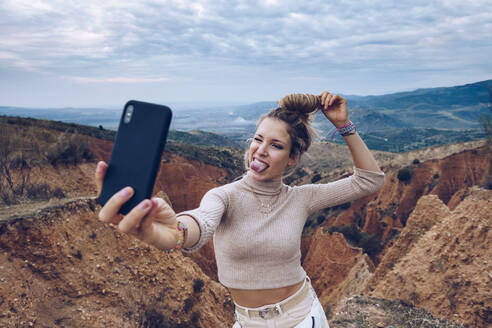 Fröhliche junge Touristin, die ein Selfie mit ihrem Smartphone macht, während sie zwischen felsigen Sandsteinformationen im Hochland steht - ADSF20524