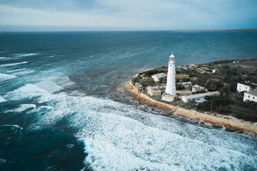 Drohnenblick von oben auf den weißen Leuchtturm und die kleine Siedlung an der felsigen Küste, die von schäumenden Meereswellen umspült wird - ADSF20529