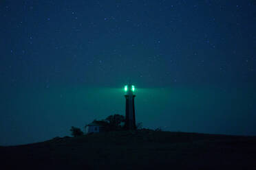 Beacon tower located on hill and illuminating darkness with green light on background of starry night sky - ADSF20531