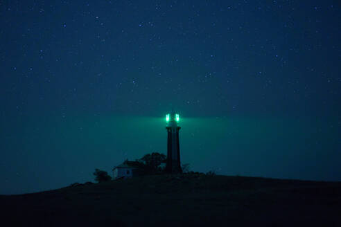 Bakenturm auf einem Hügel, der die Dunkelheit mit grünem Licht vor dem Hintergrund des nächtlichen Sternenhimmels erhellt - ADSF20531