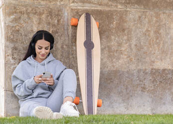 Fröhliche junge Frau in trendiger Activewear und Kopfhörern, die mit ihrem Handy telefoniert, während sie mit ihrem Longboard in der Nähe einer Steinmauer auf der Straße sitzt - ADSF20535