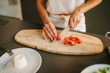 Unrecognizable female cutting ripe cherry tomatoes on wooden chopping board while standing in kitchen and cooking lunch - ADSF20537