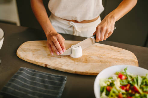 Unkenntlich erfreute Frau, die Feta-Käse auf einem Schneidebrett schneidet, während sie Zutaten für einen köstlichen hausgemachten Gemüsesalat vorbereitet - ADSF20538