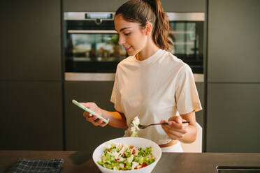 Friedliche Frau, die am Tisch in der Küche sitzt und leckeren Gemüsesalat isst, während sie auf ihrem Smartphone Nachrichten in den sozialen Medien liest - ADSF20542