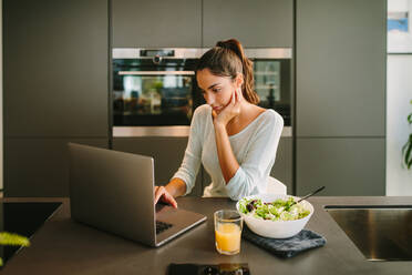 Female freelancer using laptop and working on project online while sitting at table with glass of juice and bowl of salad in kitchen - ADSF20548