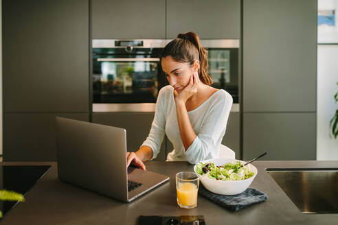 Freiberuflerin, die einen Laptop benutzt und online an einem Projekt arbeitet, während sie am Tisch mit einem Glas Saft und einer Schüssel Salat in der Küche sitzt - ADSF20548