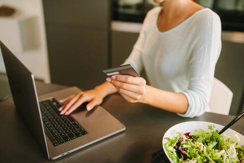 Unbekannte junge Frau mit Kreditkarte in der Hand, die einen Laptop benutzt und eine Online-Bestellung in einem Internet-Shop aufgibt, während sie in einer modernen Küche zu Hause sitzt - ADSF20549