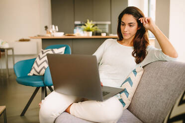Junge Freiberuflerin in legerer Kleidung, die auf dem Sofa sitzt und einen Laptop benutzt, während sie von zu Hause aus im Wohnzimmer arbeitet - ADSF20550