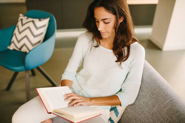 High angle of young female sitting on couch and enjoying interesting book while resting at home during weekend - ADSF20552