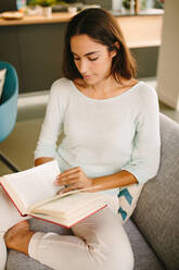 High angle of young female sitting on couch and enjoying interesting book while resting at home during weekend - ADSF20553