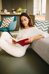 Side view of young female sitting on the floor and enjoying interesting book while resting at home during weekend - ADSF20554