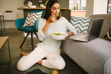 Young female in casual outfit sitting on floor and watching video on laptop while having healthy lunch with salad and cup of juice at home - ADSF20556