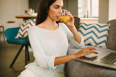 Young female drinking fresh natural orange juice while sitting on floor near sofa and browsing laptop during weekend morning at home - ADSF20557