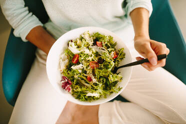 Draufsicht auf eine nicht erkennbare junge Frau in Freizeitkleidung, die auf einem Stuhl sitzt und einen gesunden vegetarischen Salat in einer modernen Wohnküche isst - ADSF20558