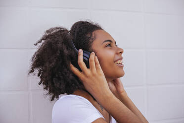 Seitenansicht einer zufriedenen afroamerikanischen Frau mit kabellosen Kopfhörern, die auf der Straße steht und nach oben schaut, während sie ein Lied hört - ADSF20559