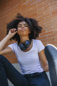 Positive afroamerikanische Frau mit lockigem Haar, die mit Kopfhörern und Skateboard auf der Straße sitzt, während sie in die Kamera schaut und das Wochenende in der Stadt genießt - ADSF20560