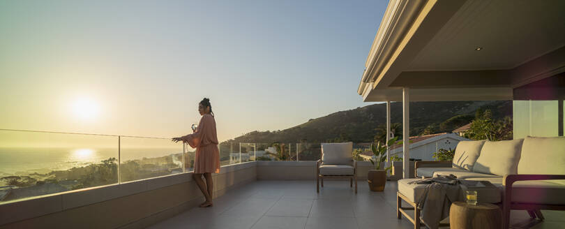 Frau mit Weißwein genießt den Sonnenuntergang mit Blick auf den Ozean auf einem Luxusbalkon - CAIF30229