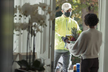 Woman receiving grocery delivery from courier in face mask at door - CAIF30262