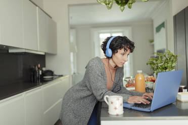 Frau mit Kopfhörern arbeitet von zu Hause aus am Laptop in der Küche - CAIF30271