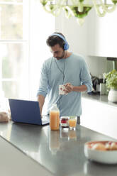 Mann mit Kopfhörern arbeitet von zu Hause aus am Laptop in der Morgenküche - CAIF30276